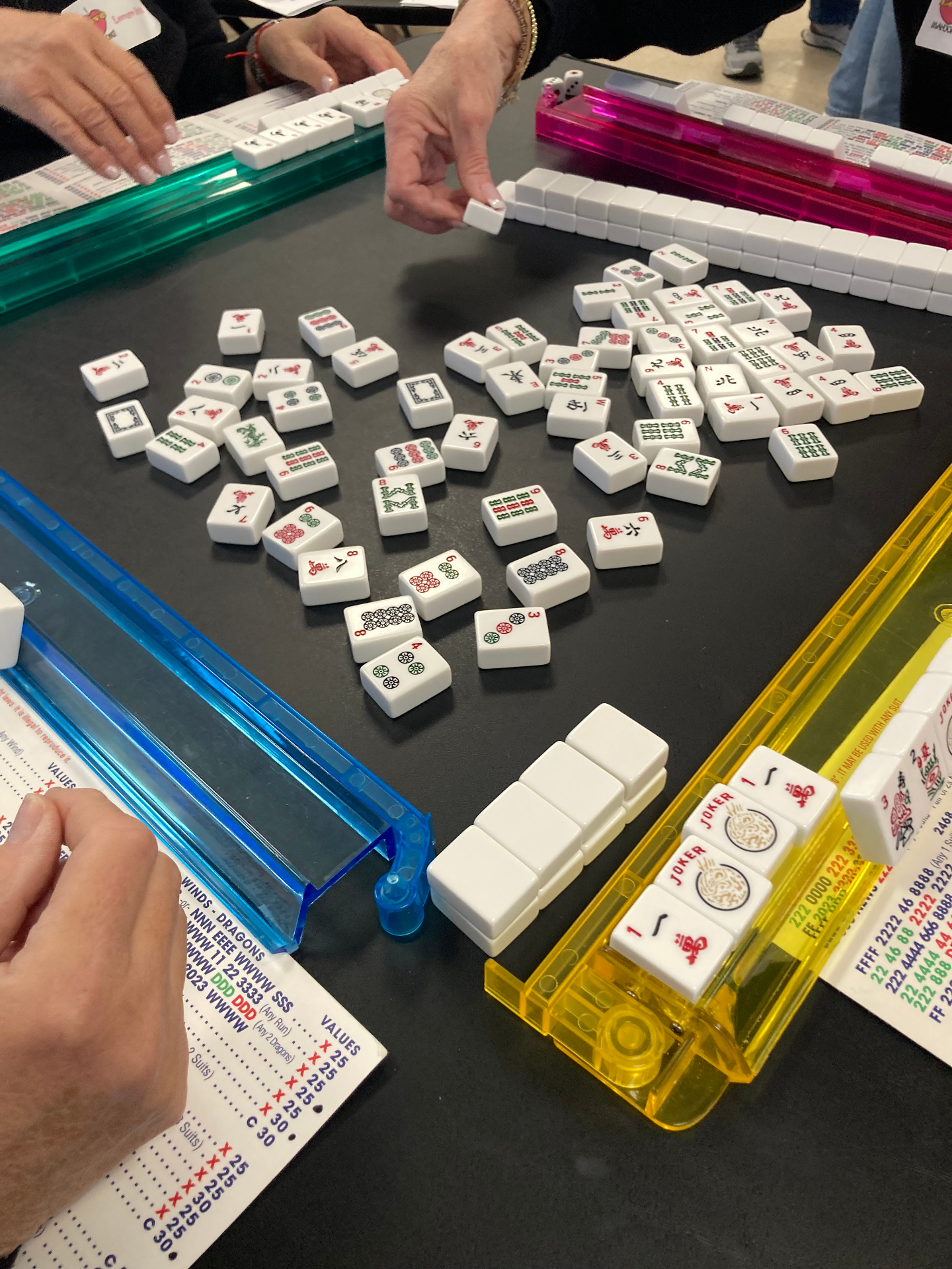 Players around a Mah Jongg table mid-game with tiles, racks, and score cards